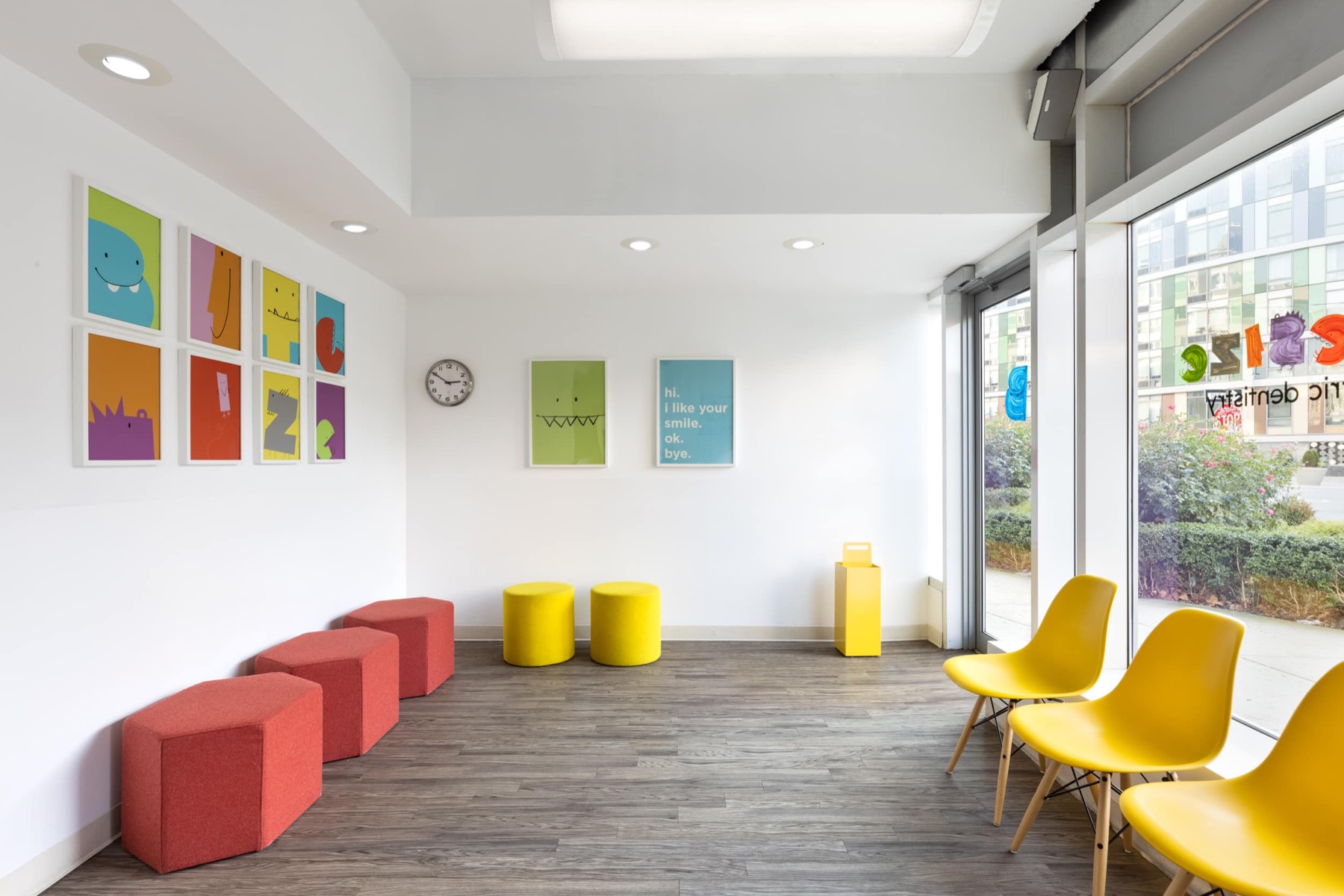 Wide view of the Bitesize Williamsburg waiting room with the poster grid, a wall clock and the teal hi i like your smile ok bye poster, daylight through the shopfront glazing