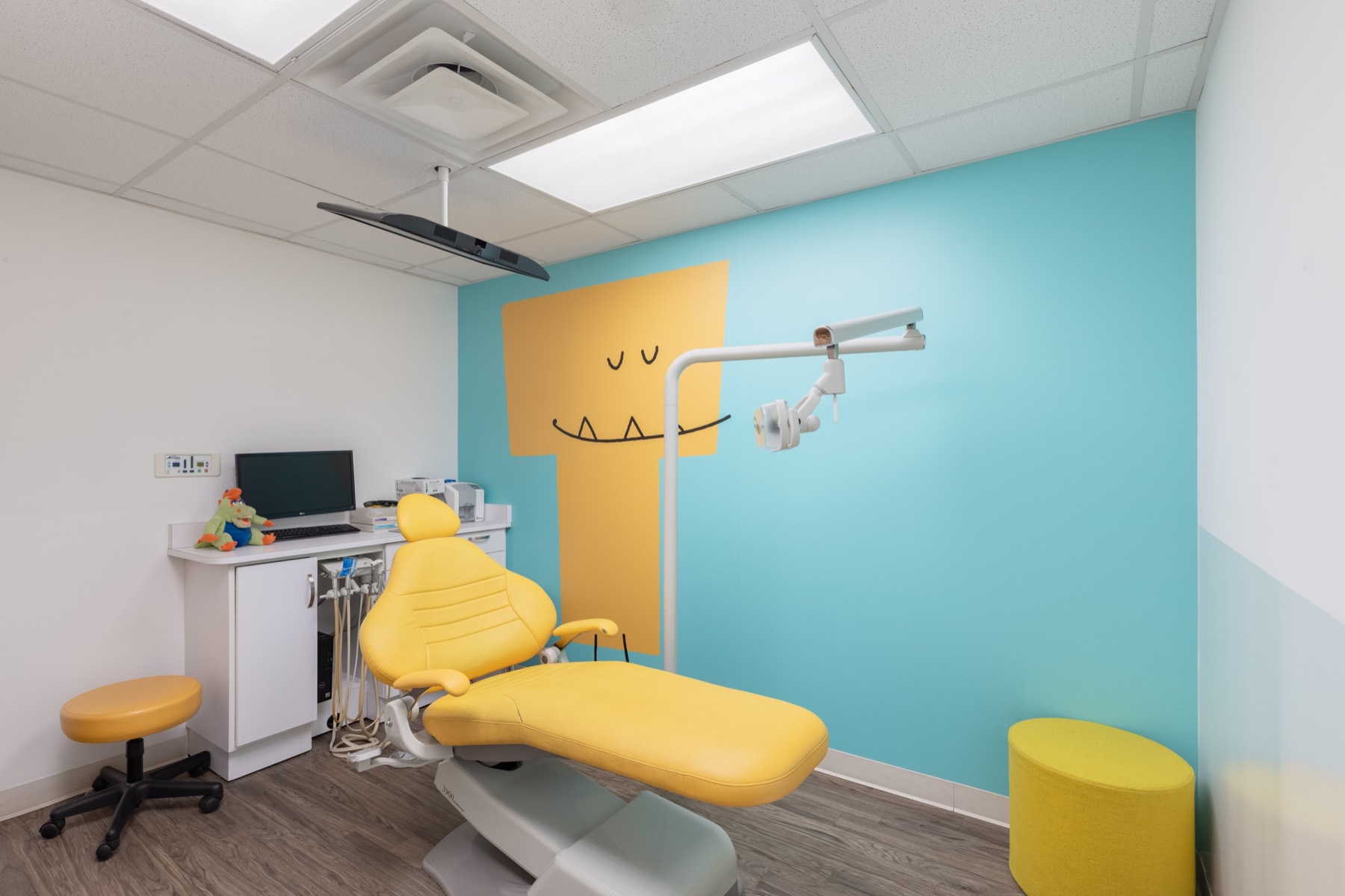 A treatment room at Bitesize Williamsburg with a yellow dental chair, a painted yellow monster mural on a pale blue wall and a flatscreen TV mounted on the ceiling above the chair